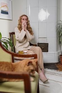A woman sneezing in a cozy interior with a ginger cat and houseplants, depicting allergy season.