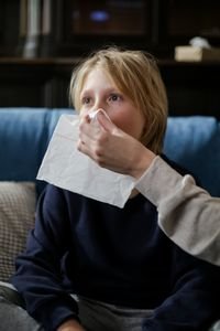 A young child holding a tissue, assisted by an adult, for sneezing or illness recovery indoors.